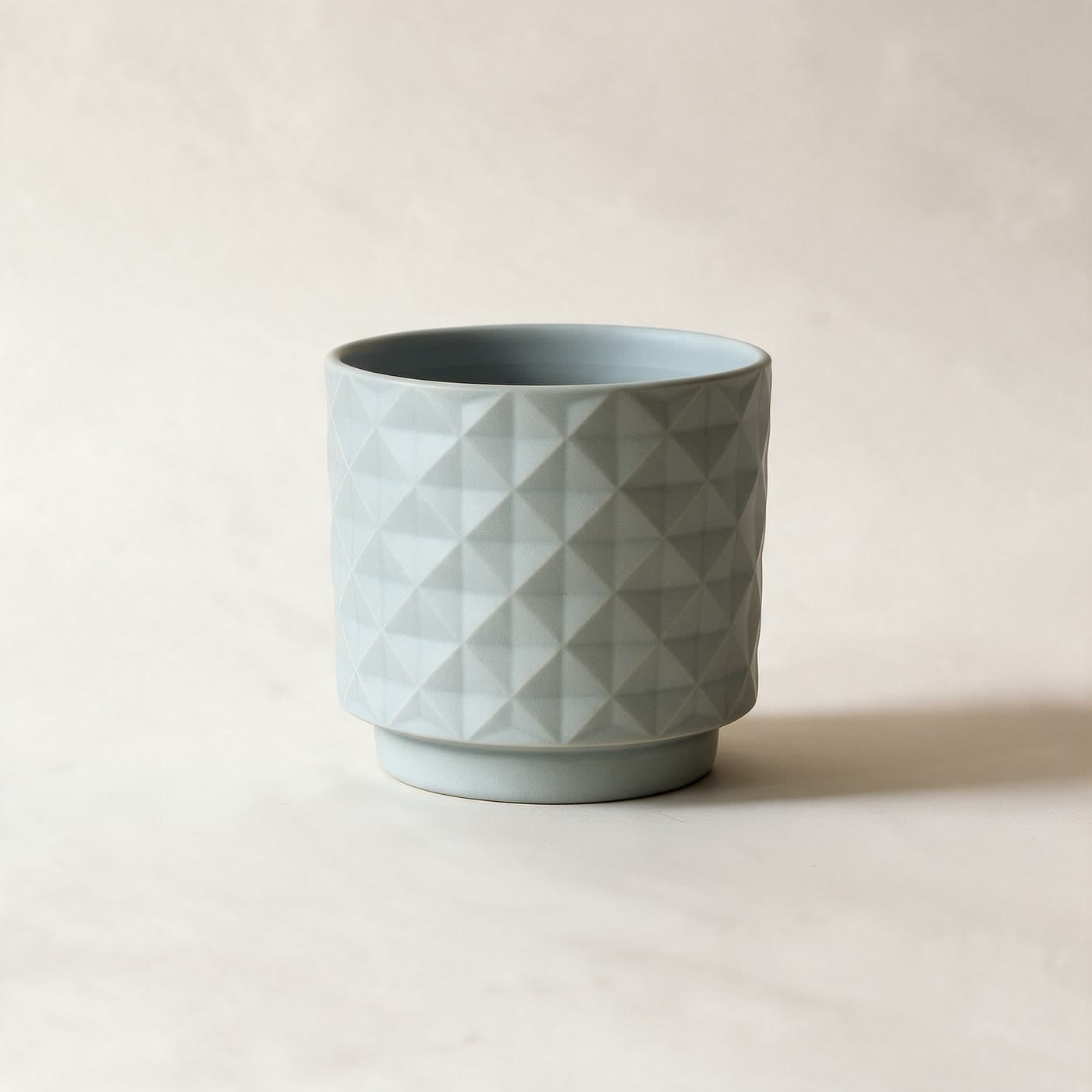 Small matte grey-blue ceramic planter with an all-over raised diamond facet texture and a small attached saucer foot, standing alone on a plain warm off-white studio background