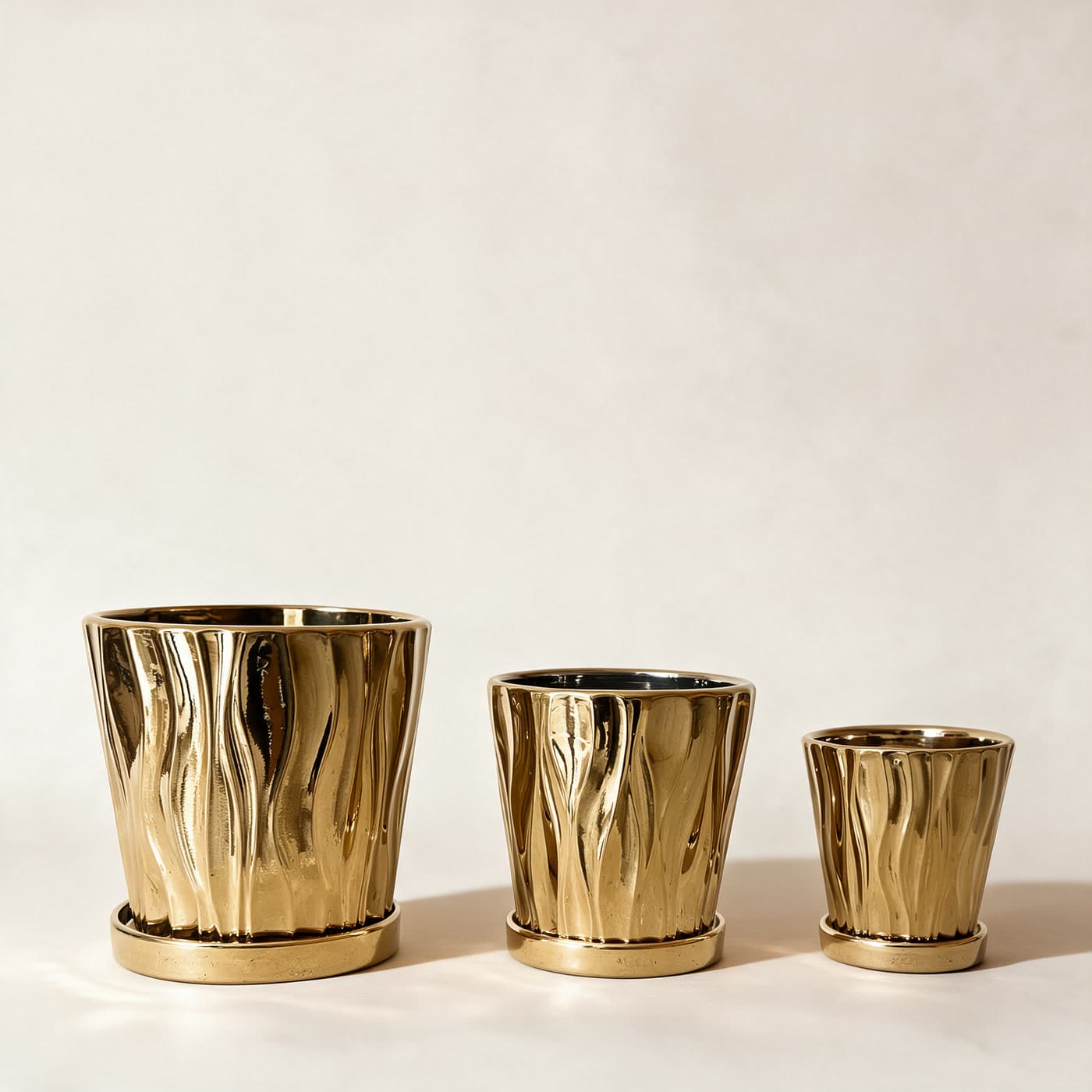 Three mirror-gold ceramic planters in a row, large to small, each moulded in tall wavering flutes and standing on its matching gold saucer, on a plain warm off-white studio background