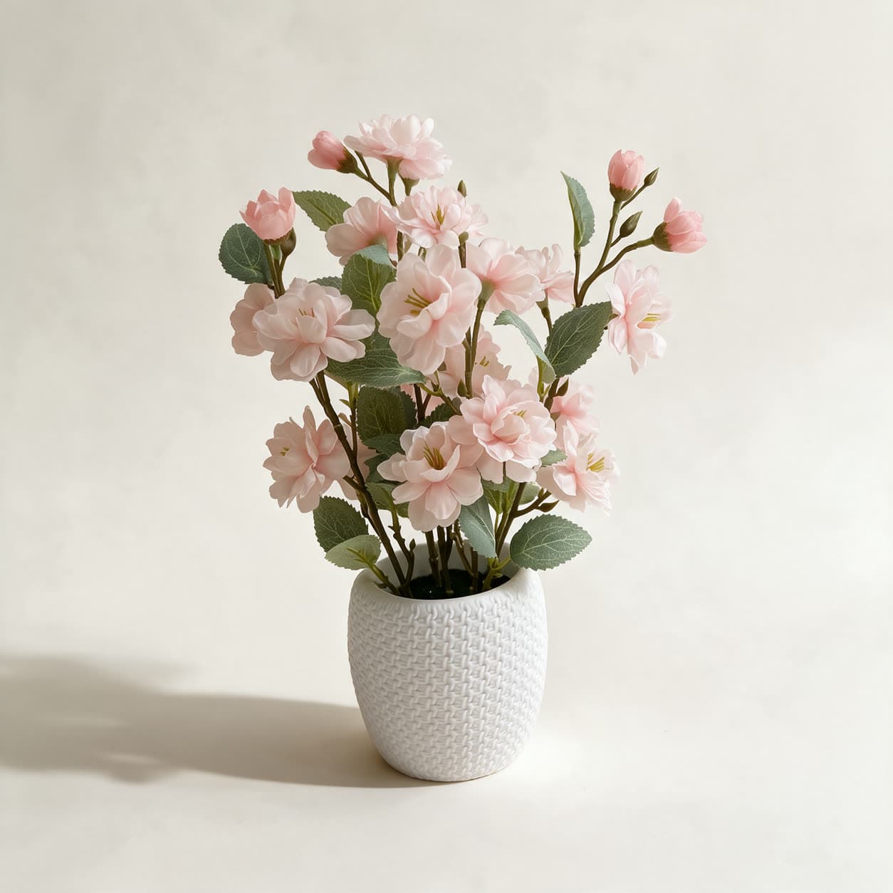 Small artificial arrangement of soft pink blossoms in a white basketweave-textured ceramic pot, on a warm off-white background