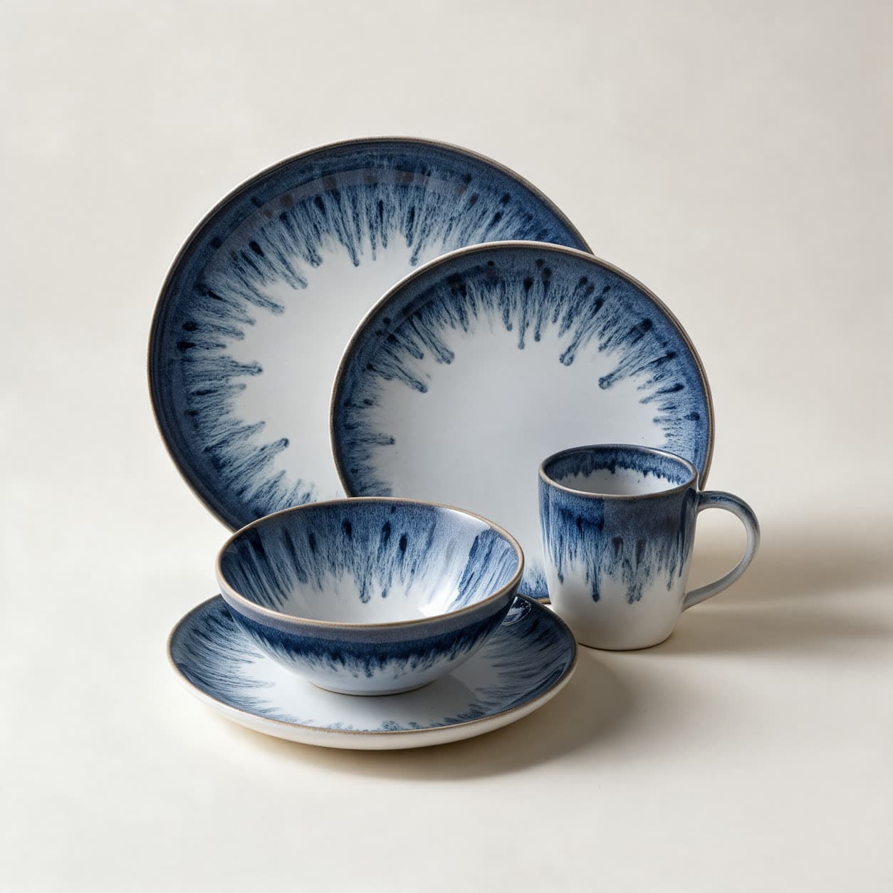 Indigo and white reactive-glaze stoneware place setting — dinner plate, side plate, bowl on a saucer and a mug — with deep blue feathering down from the rims, on a warm off-white background