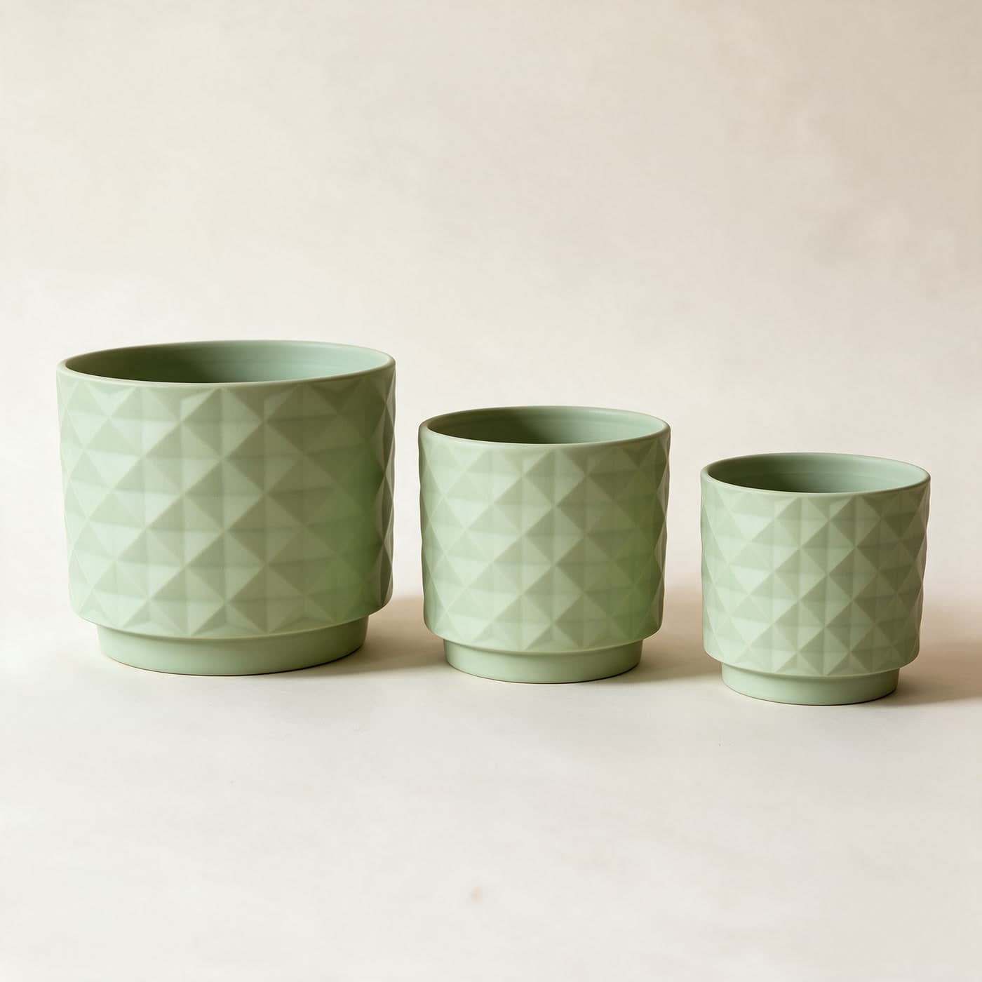 Three matte sage green ceramic planters in a row, large to small, each with an all-over raised diamond facet texture and a small attached saucer foot, on a plain warm off-white studio background