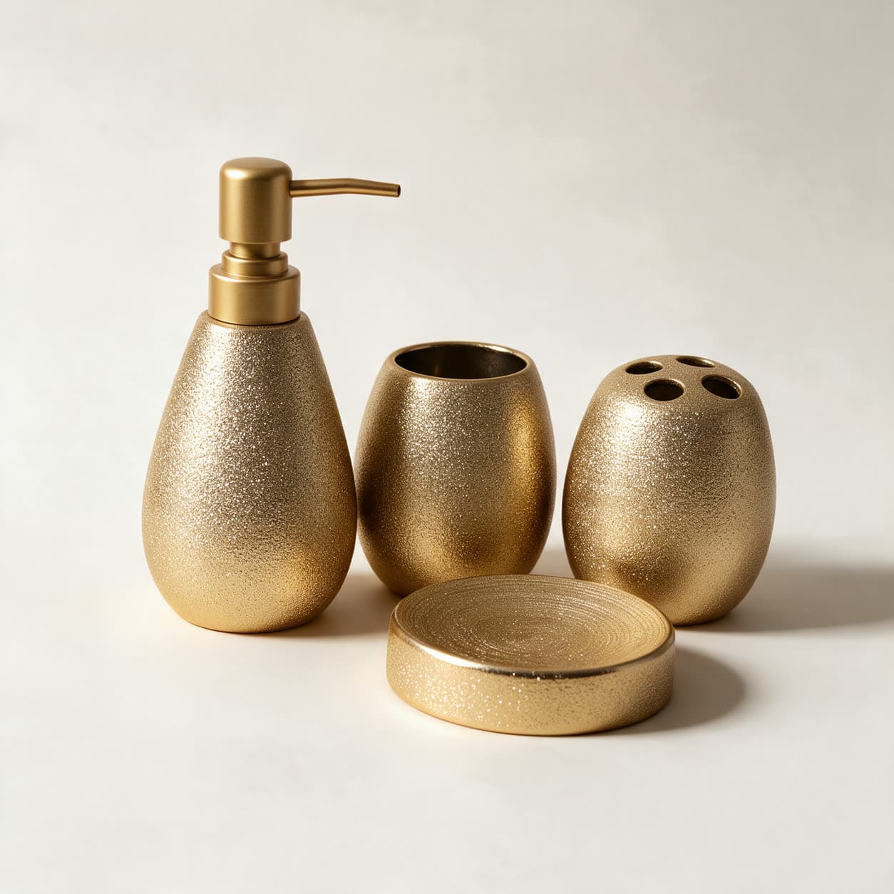 Four-piece bathroom set in textured sparkling gold — a teardrop soap dispenser, an egg-shaped tumbler, a toothbrush holder with five holes and a round soap dish — on a warm off-white background