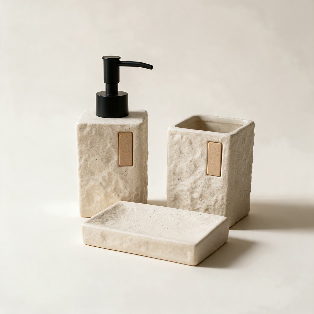 Three-piece cream stone-look bathroom set — a square soap dispenser with a black pump, a square tumbler and a rectangular soap dish — on a warm off-white background