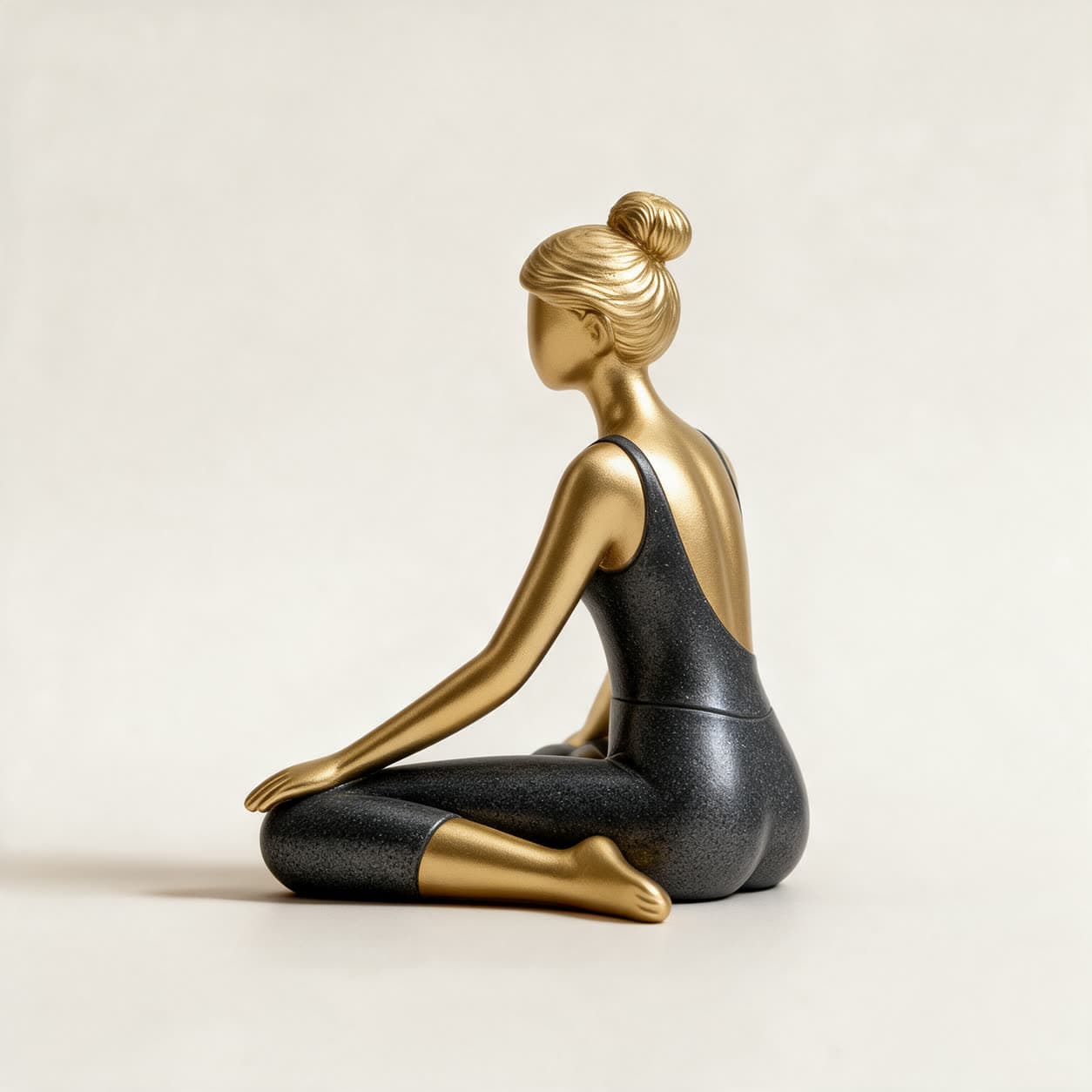 Small cast resin figurine of a woman seated in a yoga stretch, with polished gold skin and hair and a dark charcoal leotard, on a warm off-white background