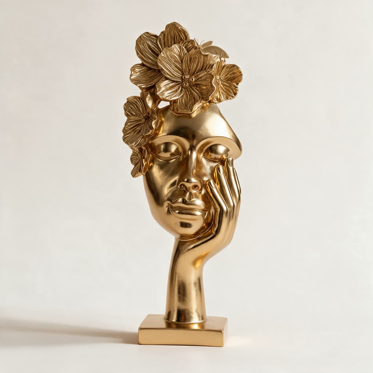 Polished gold cast resin sculpture of a stylised face with a crown of sculpted flowers and a hand resting against the cheek, on a square base, on a warm off-white background