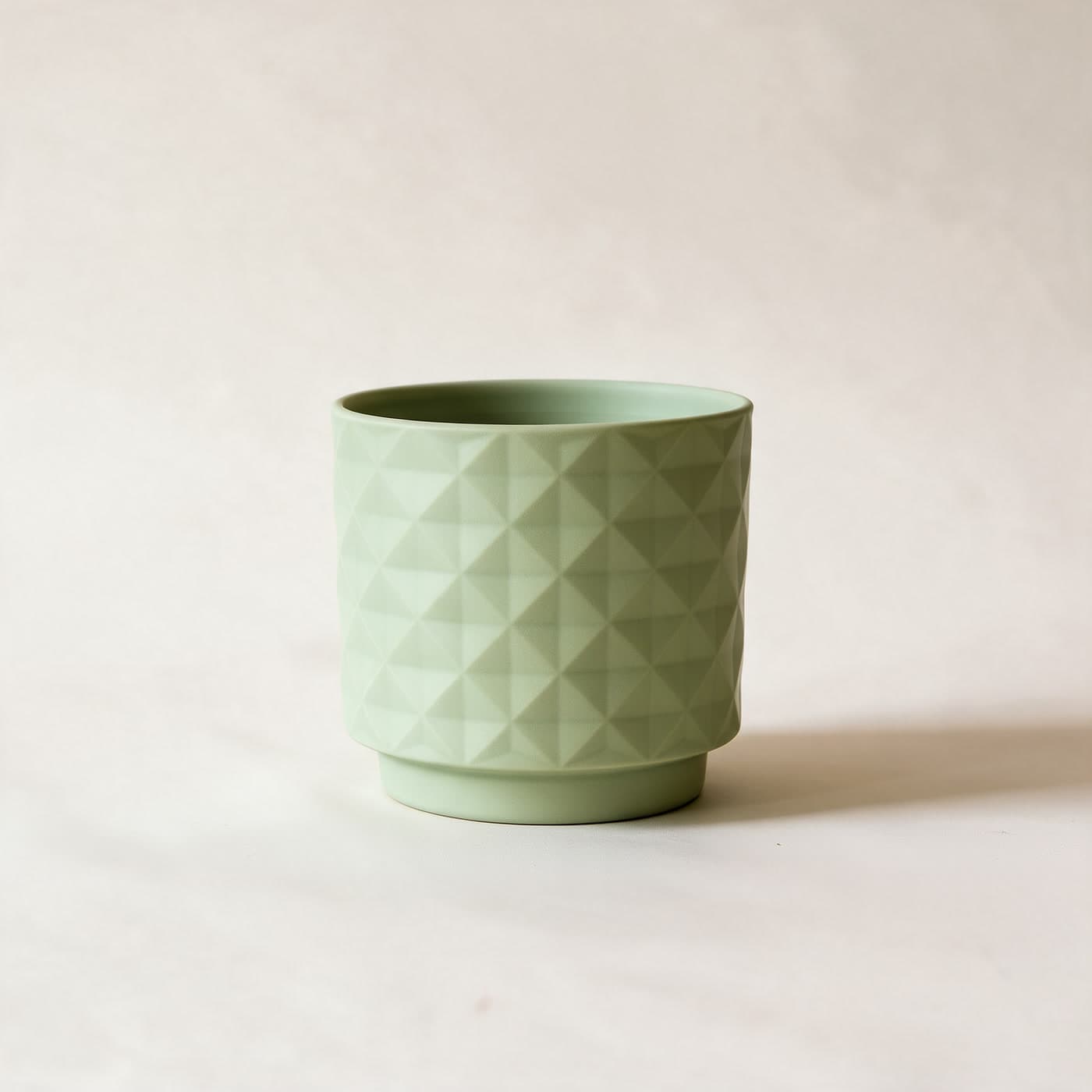Small matte sage green ceramic planter with an all-over raised diamond facet texture and a small attached saucer foot, standing alone on a plain warm off-white studio background