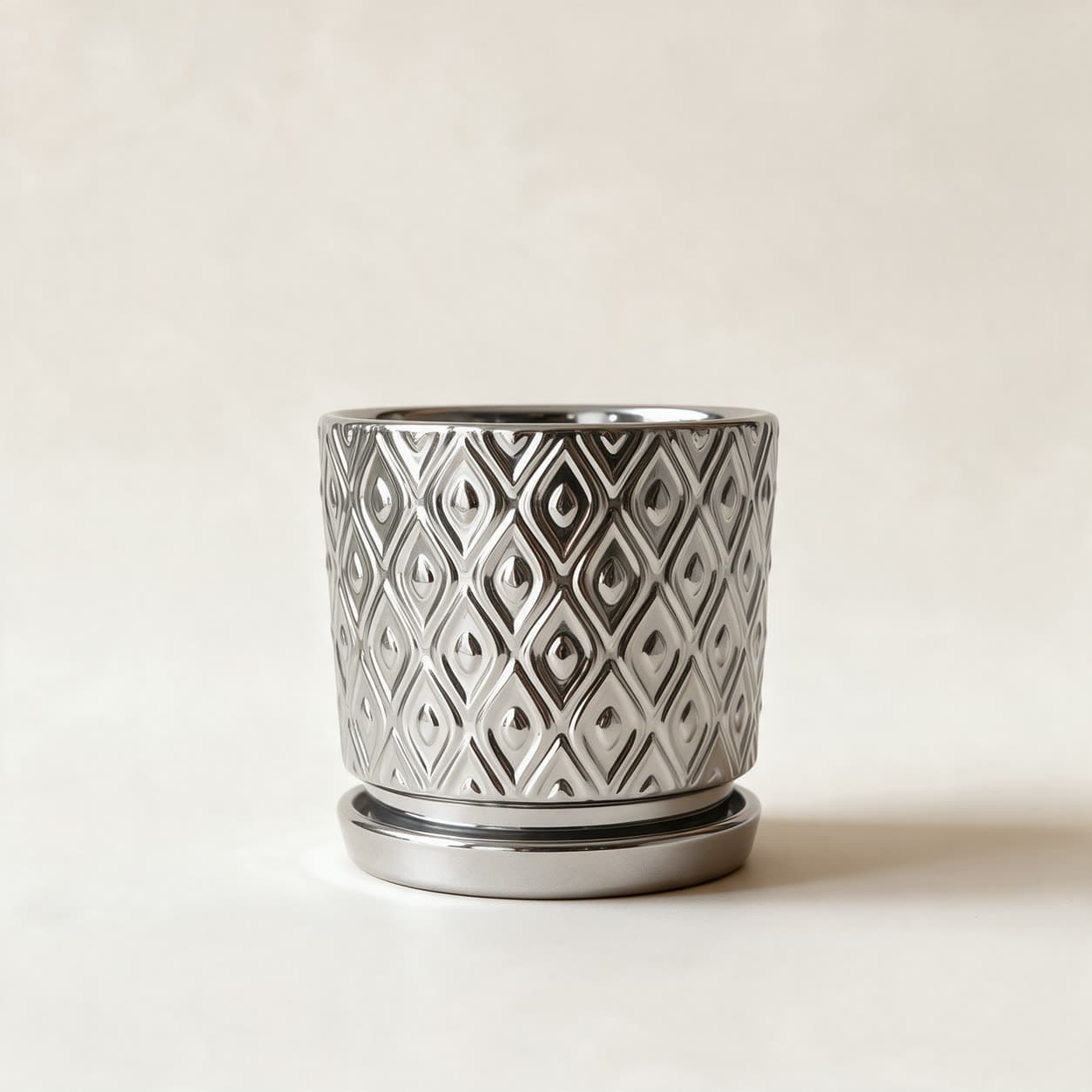 Cylindrical ceramic planter in mirror-silver with an embossed lattice of pointed ovals, sitting on a matching silver saucer, on a warm off-white background