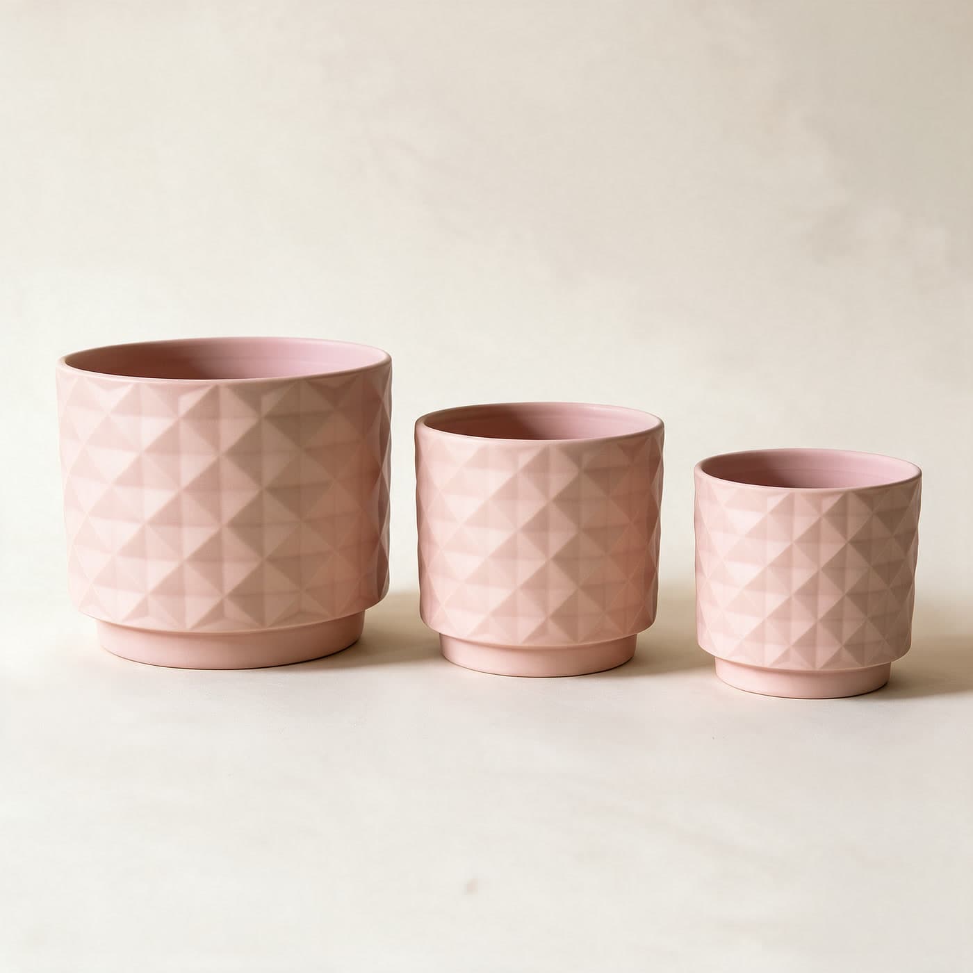 Three matte blush pink ceramic planters in a row, large to small, each with an all-over raised diamond facet texture and a small attached saucer foot, on a plain warm off-white studio background