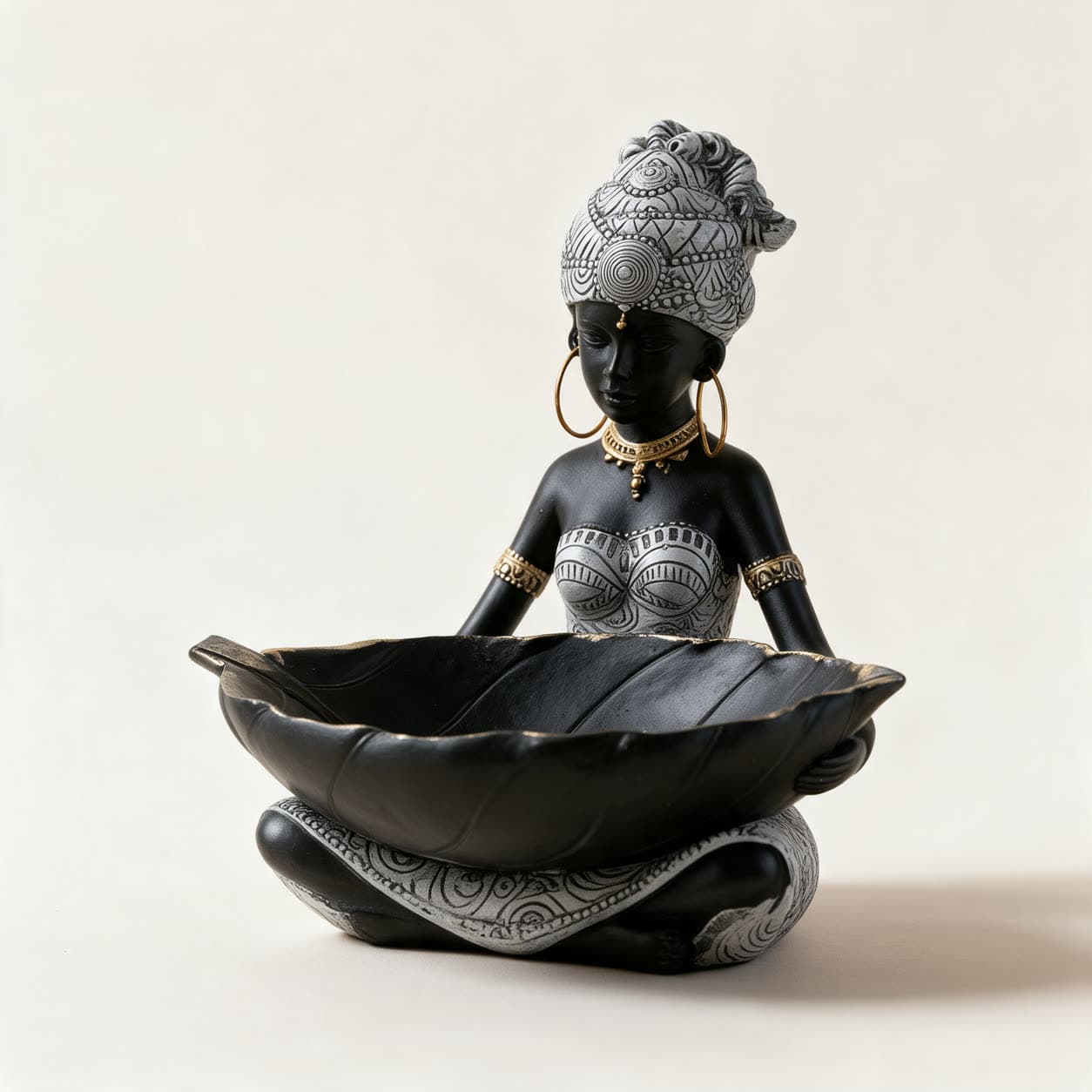 Cast resin figurine of a seated woman in matte black with a patterned headwrap and gold jewellery, holding a large black leaf-shaped dish edged in gold, on a warm off-white background