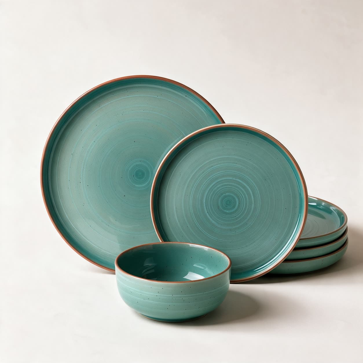 Turquoise speckled stoneware dinner set with rust-brown unglazed rims — dinner plate, side plate, bowl and a stack of further plates — on a warm off-white background