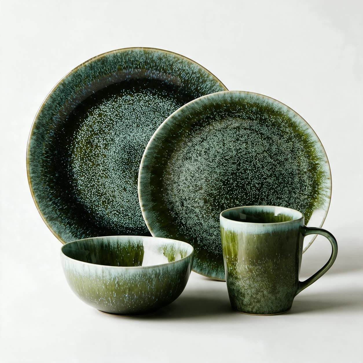 Four-piece forest green reactive-glaze stoneware place setting — dinner plate, side plate, bowl and mug — speckled with cream and breaking lighter at the rims, on a warm off-white background