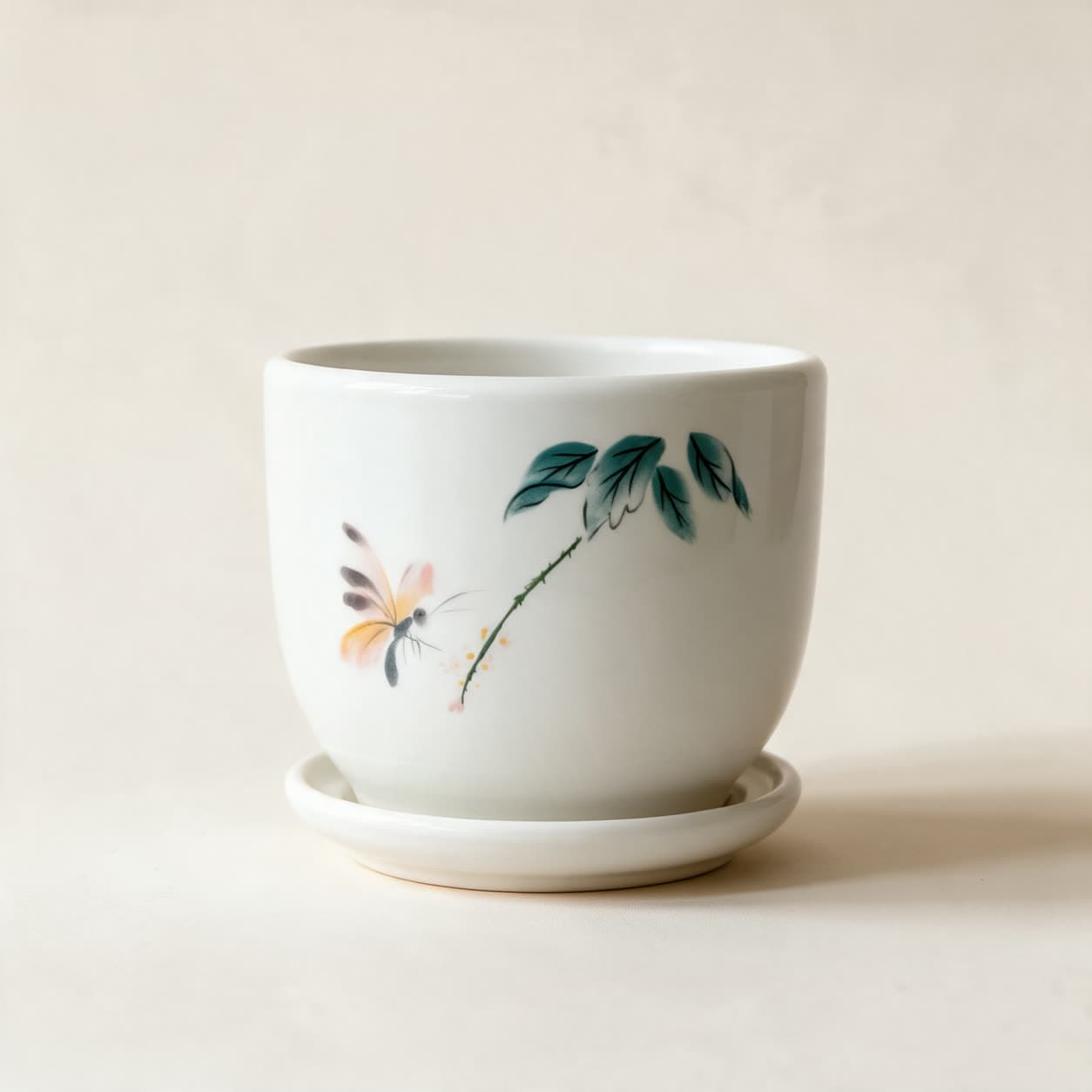 Rounded glossy white ceramic planter hand-painted with a teal leafy stem and a soft orange butterfly, on a matching white saucer, on a warm off-white background