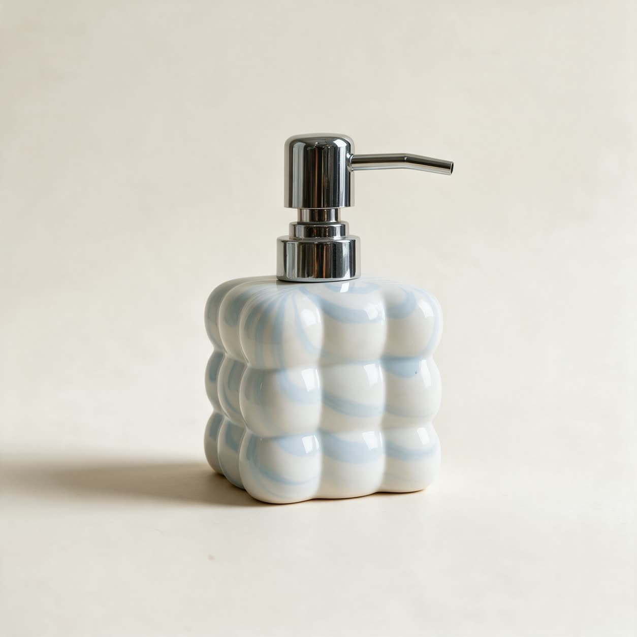 Square cloud-shaped soap dispenser in pale blue and white swirled ceramic with a chrome pump, on a warm off-white background