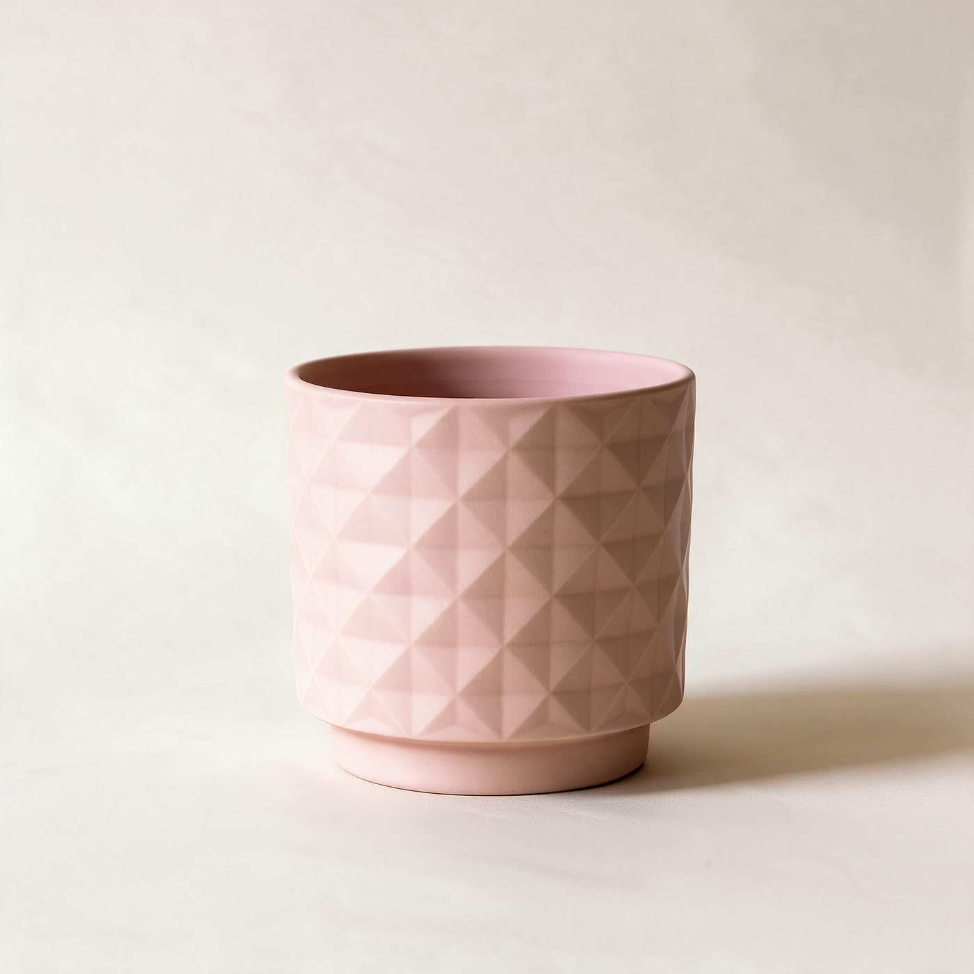 Small matte blush pink ceramic planter with an all-over raised diamond facet texture and a small attached saucer foot, standing alone on a plain warm off-white studio background