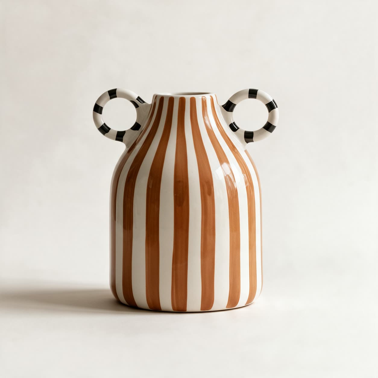 Glossy ceramic vase hand-painted with broad terracotta and cream vertical stripes, with two open ring handles banded in black and white, on a warm off-white background