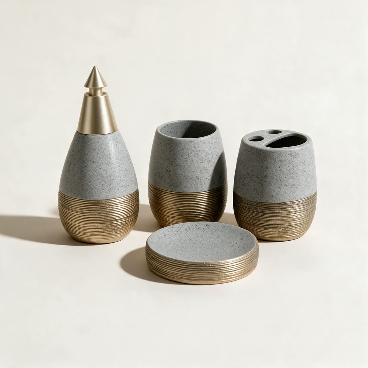 Four-piece grey speckled stone-look bathroom set with ribbed gold bands — teardrop soap dispenser, tumbler, toothbrush holder and round soap dish — on a warm off-white background