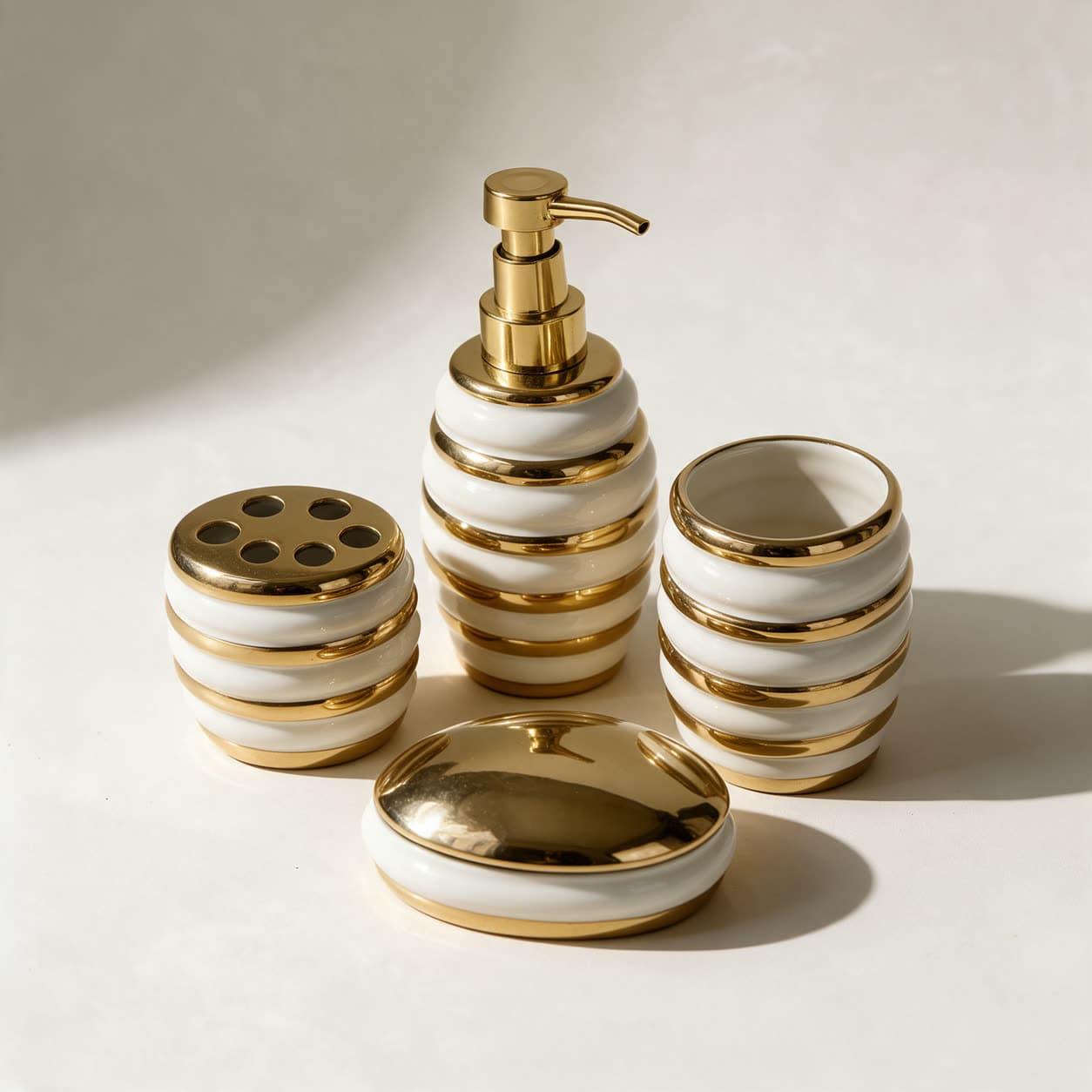 Four-piece bathroom set in glossy ivory ceramic with polished gold bands — soap dispenser, tumbler, toothbrush holder and covered soap dish — on a warm off-white background