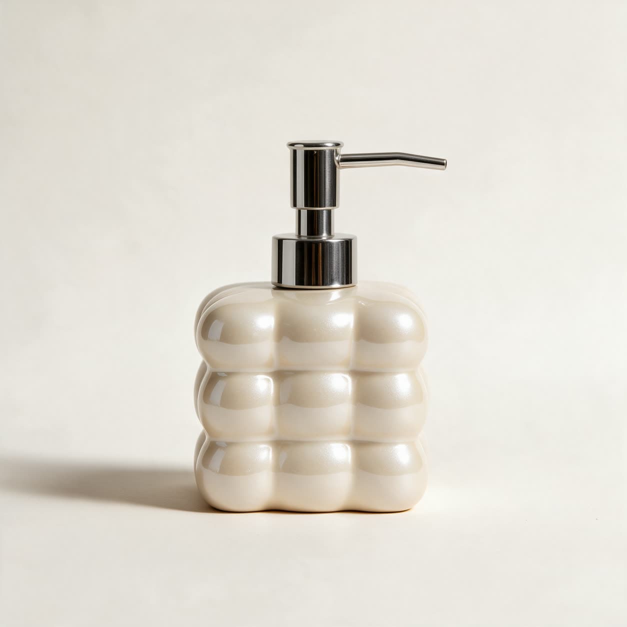 Square cloud-shaped soap dispenser in pearly ivory ceramic with a chrome pump, on a warm off-white background