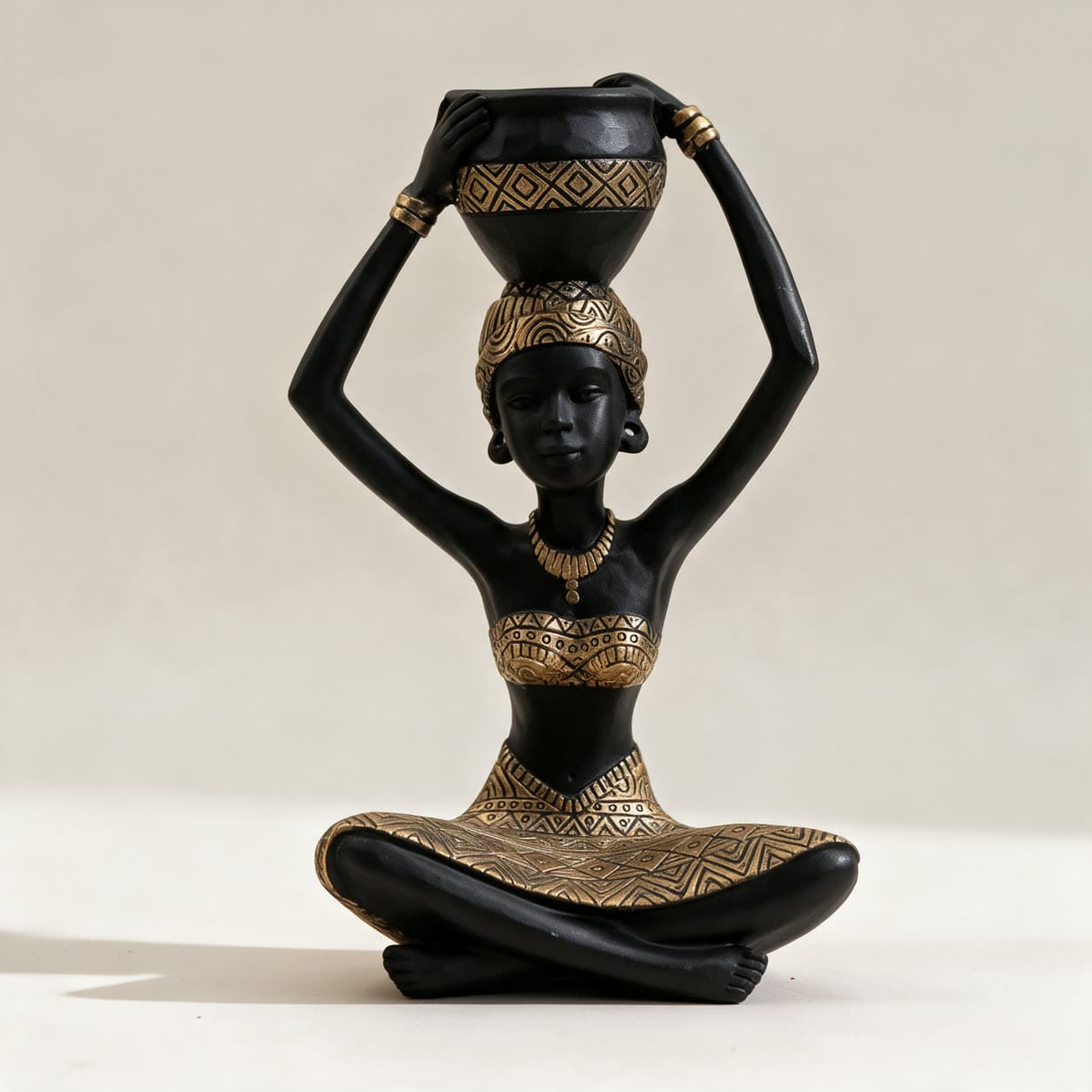 Cast resin figurine of a kneeling woman in matte black with gold geometric patterning, holding a round pot above her head, on a warm off-white background