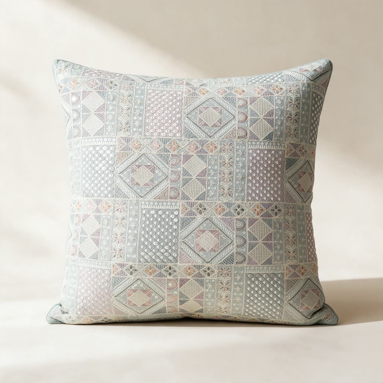 Pale sage-blue cushion cover hand-embroidered in a dense patchwork of diamonds, lattice and small flowers in cream and white with pink and lilac accents and small round mirrors, on a warm off-white background