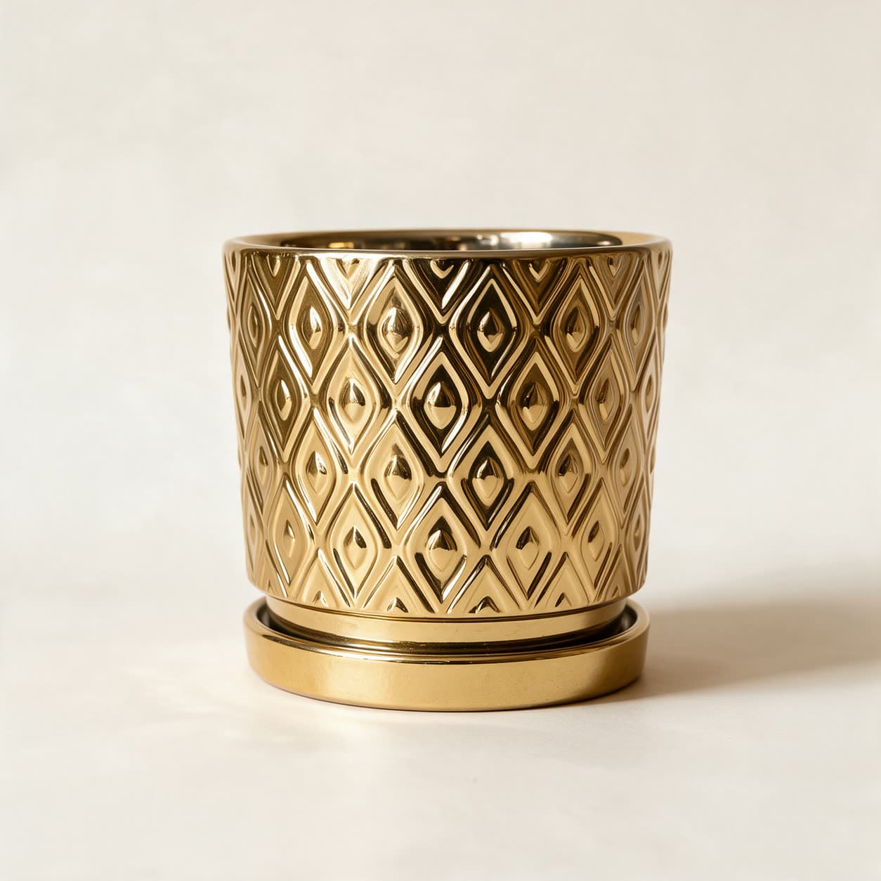 Cylindrical ceramic planter in mirror-gold with an embossed lattice of pointed ovals, sitting on a matching gold saucer, on a warm off-white background