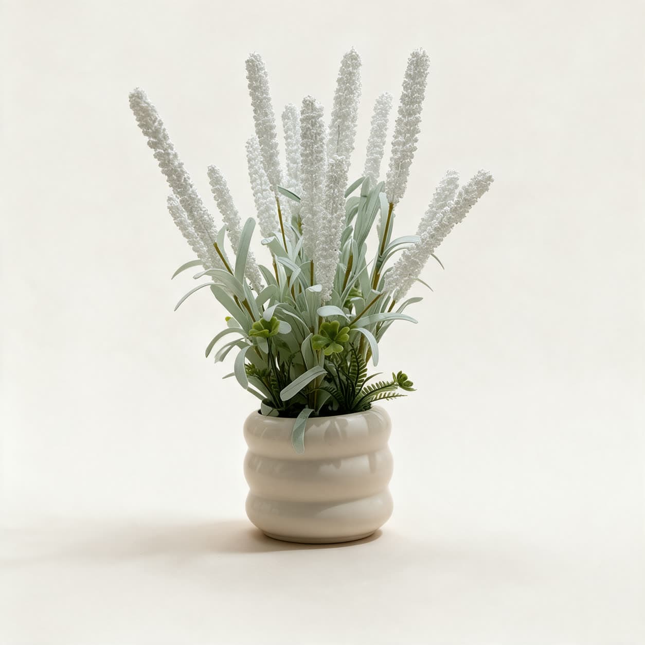 Small artificial arrangement of upright white floret spikes with grey-green leaves in a ridged cream ceramic pot, on a warm off-white background