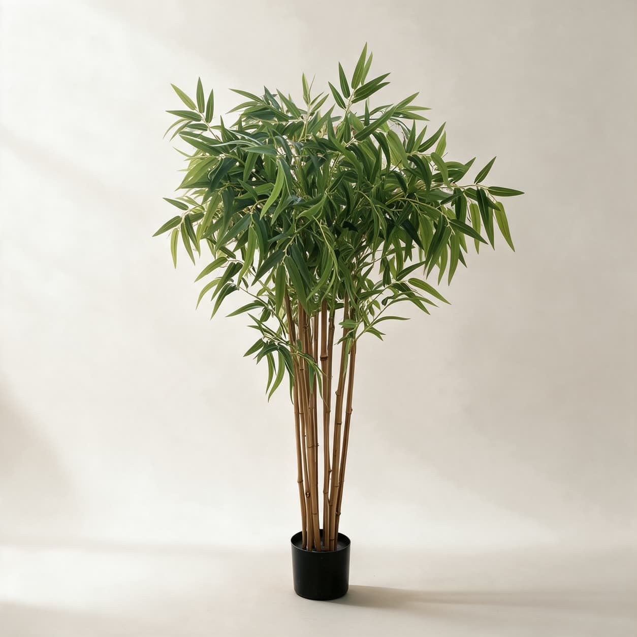 Artificial bamboo tree about 1.2 metres tall, slim tan canes with long narrow green leaves, in a black pot on a warm off-white background