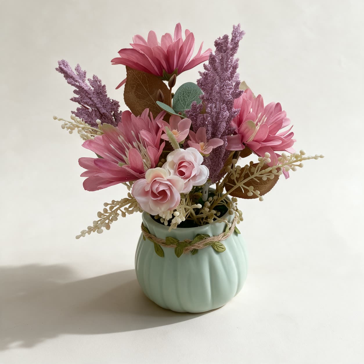 Small artificial posy of pink daisies, a pale rose, mauve spikes and cream berries in a mint-green ribbed ceramic pot with twine trim, on a warm off-white background