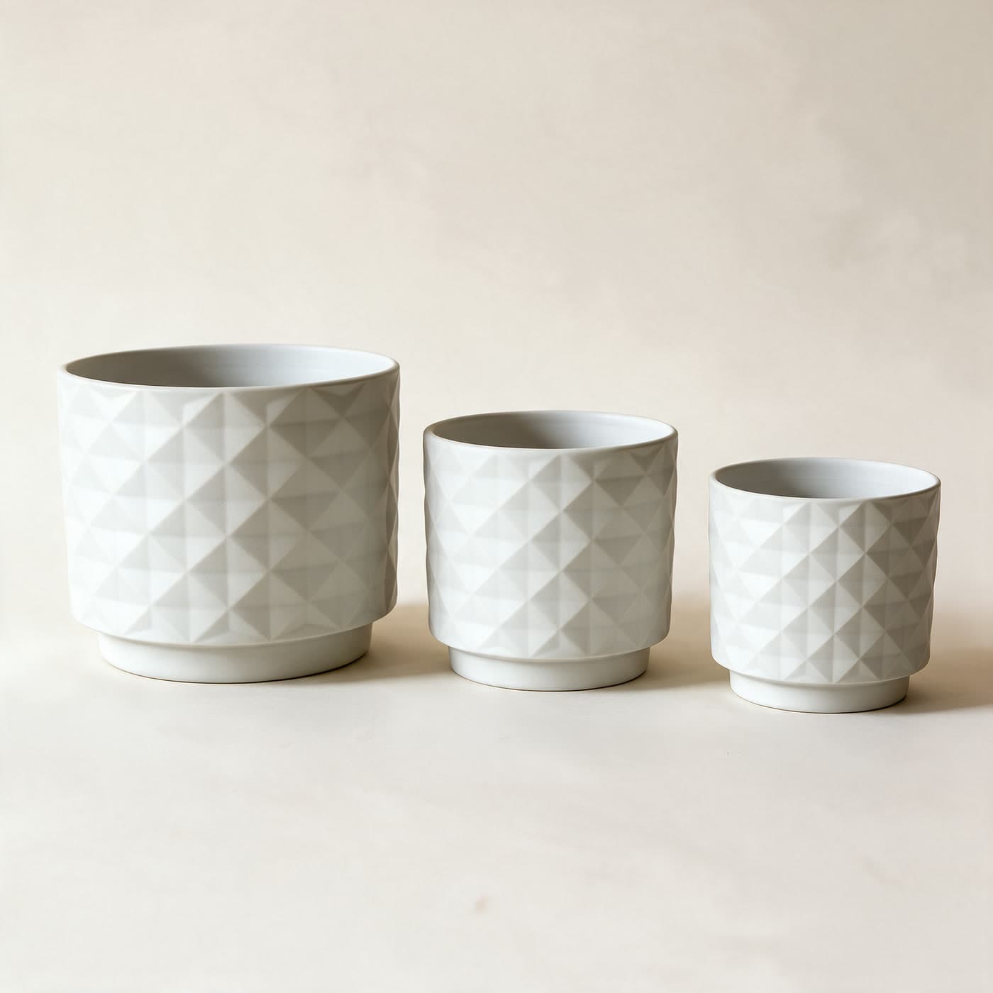 Three matte chalk white ceramic planters in a row, large to small, each with an all-over raised diamond facet texture and a small attached saucer foot, on a plain warm off-white studio background