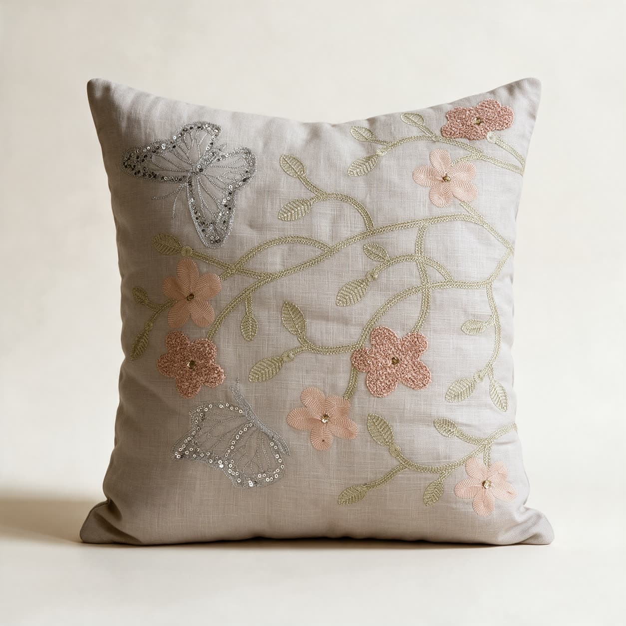 Pale stone-grey linen cushion cover hand-embroidered with a sage-gold vine, blush flowers with gold bead centres and two silver sequinned butterflies, on a warm off-white background