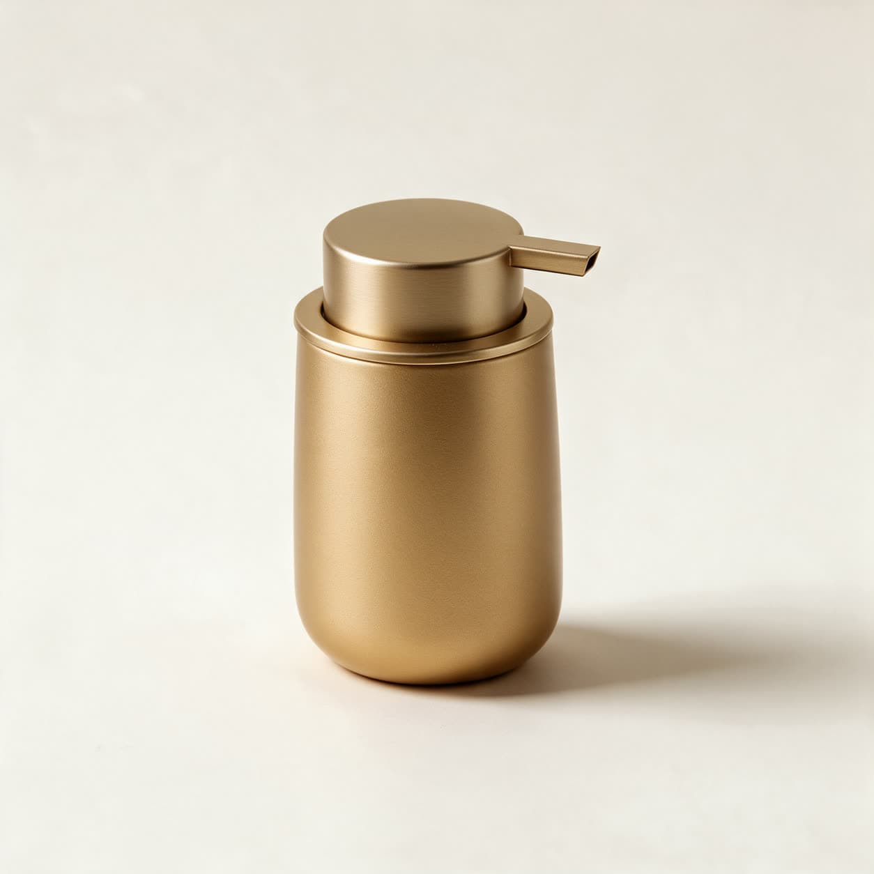 Cylindrical soap dispenser in matte gold with a flat brushed gold disc lid and a side press lever, on a warm off-white background