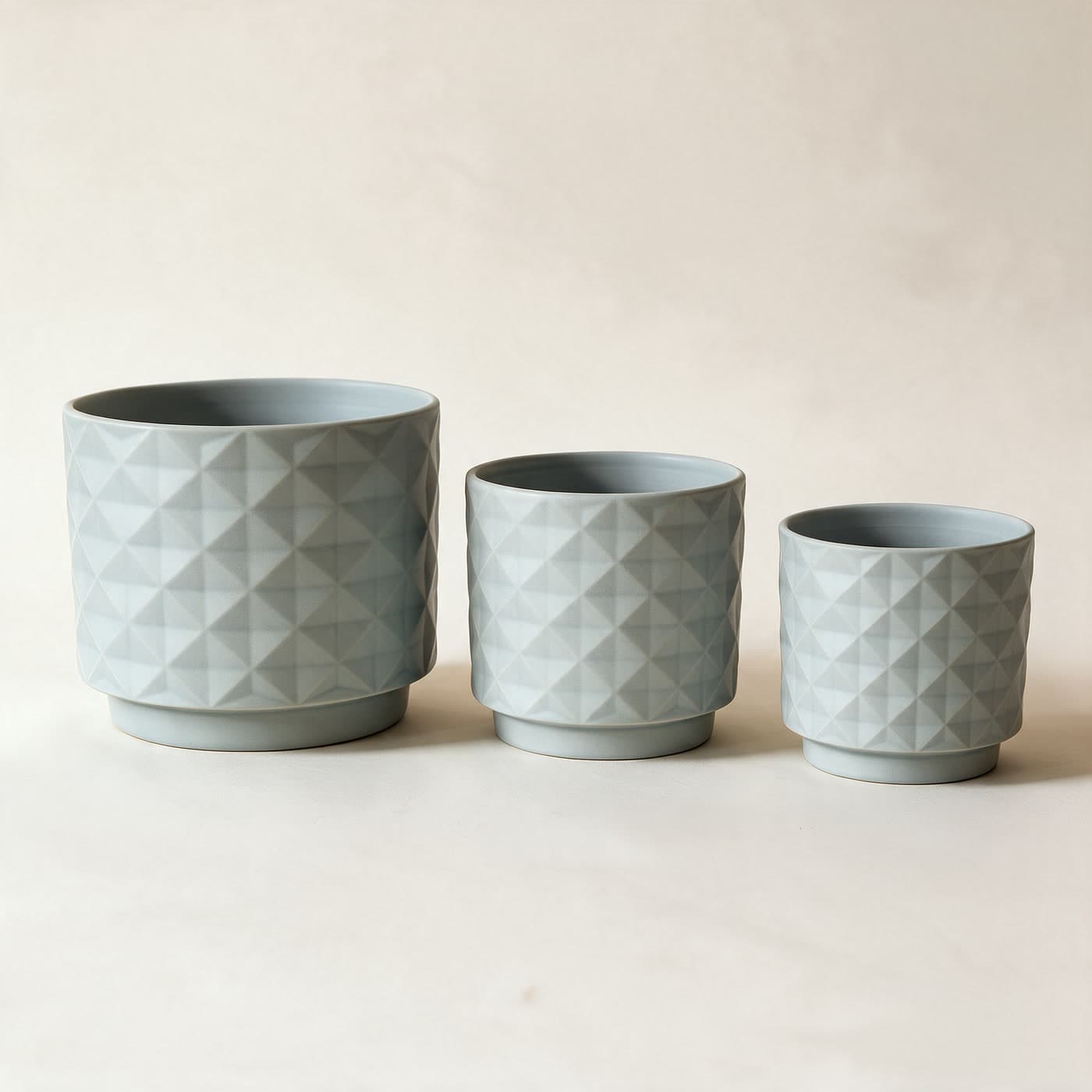Three matte grey-blue ceramic planters in a row, large to small, each with an all-over raised diamond facet texture and a small attached saucer foot, on a plain warm off-white studio background