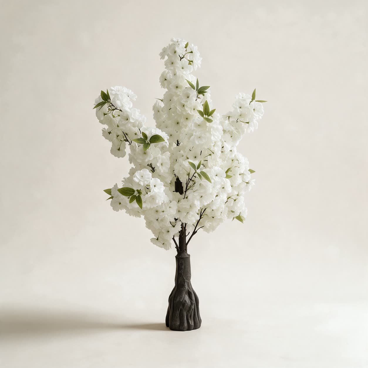 Artificial white blossom tree about 1 metre tall, branches densely covered in small white flowers, rising from a dark root-shaped base, on a warm off-white background