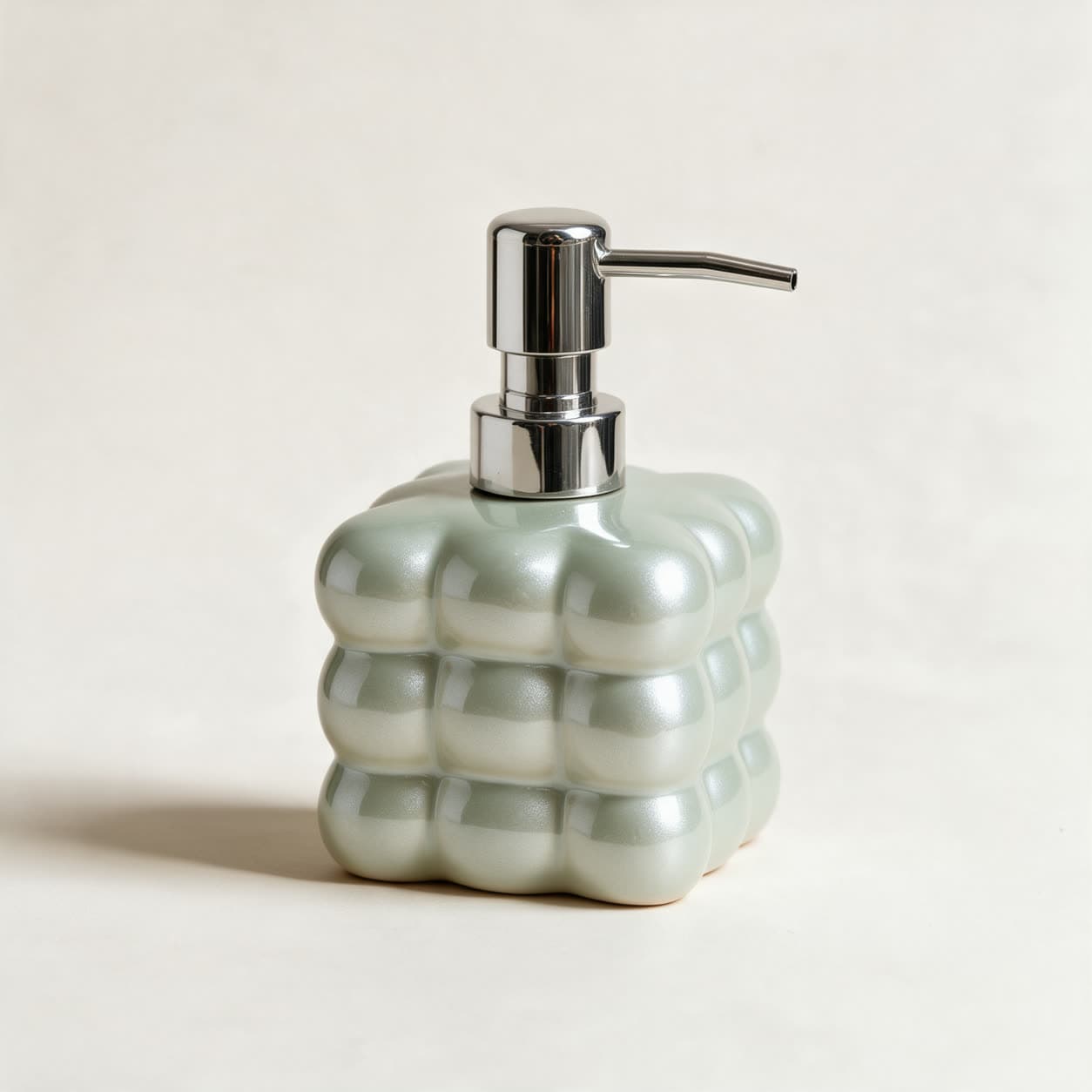 Square cloud-shaped soap dispenser in pearly sage green ceramic with a chrome pump, on a warm off-white background