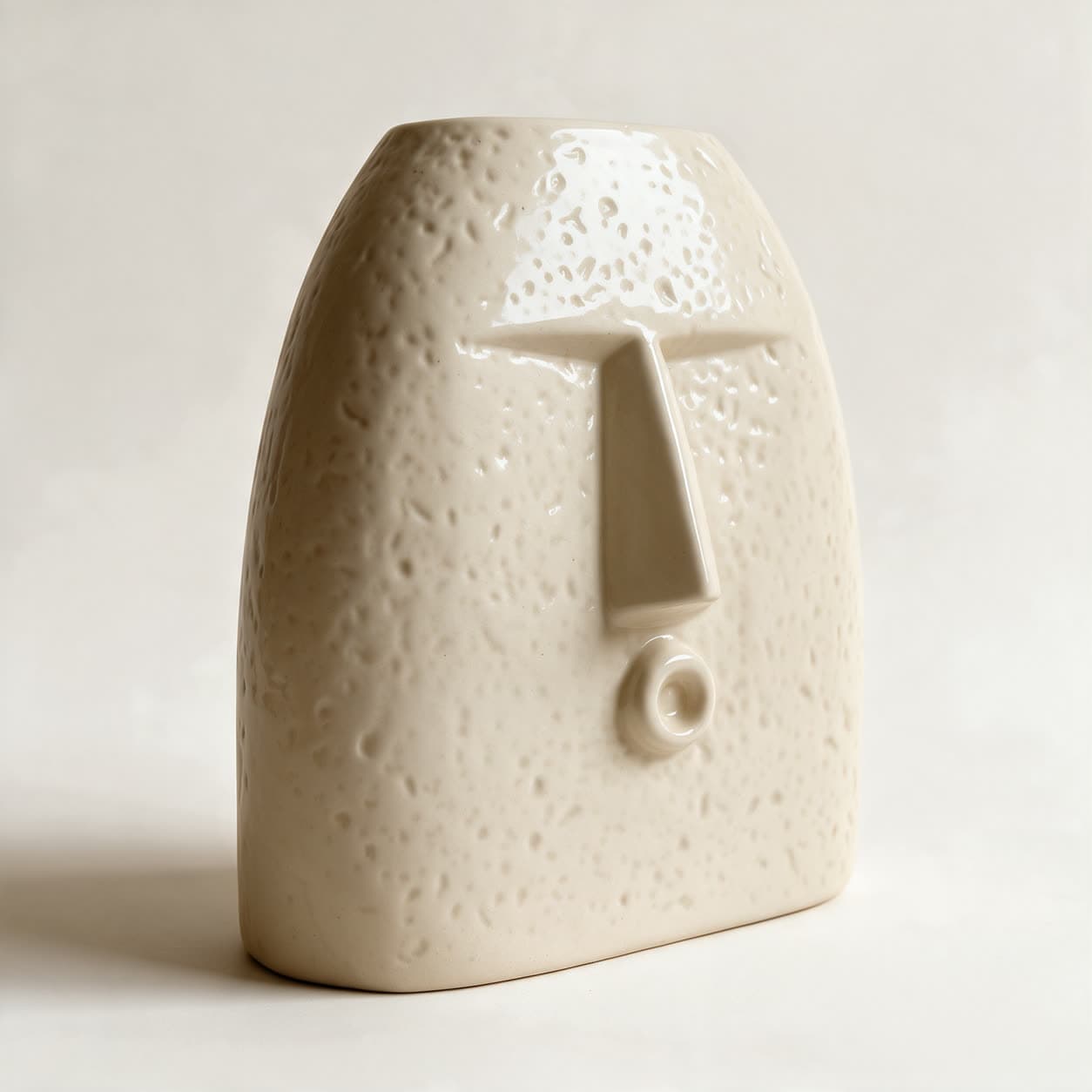 Tall ivory ceramic face planter with an abstracted face — a straight brow, a long wedge nose and one small raised ring — on a finely stippled surface, on a warm off-white background