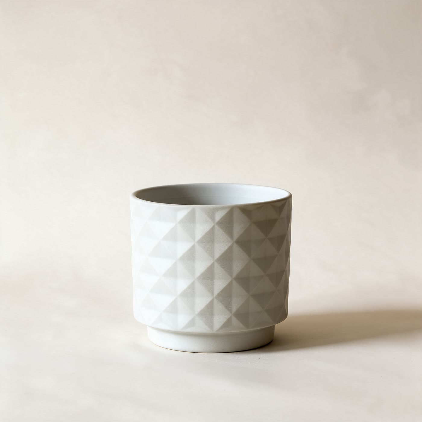 Small matte chalk white ceramic planter with an all-over raised diamond facet texture and a small attached saucer foot, standing alone on a plain warm off-white studio background