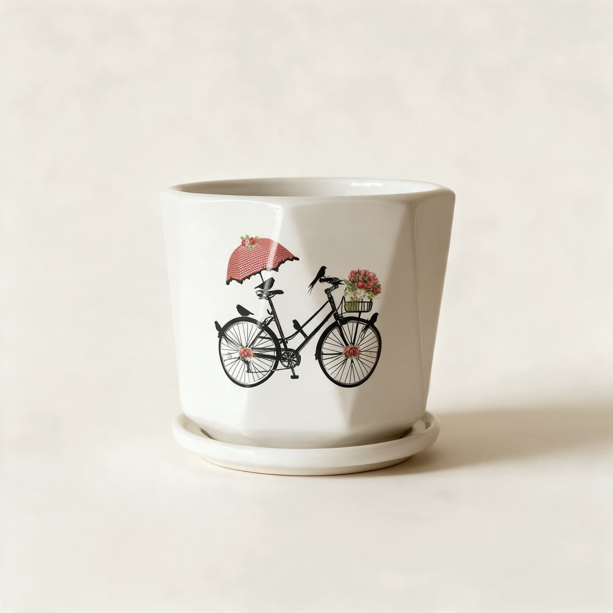 Faceted cream-white ceramic planter printed with a black line drawing of an upright bicycle carrying a basket of pink roses and an open spotted parasol, on a matching saucer, on a warm off-white background