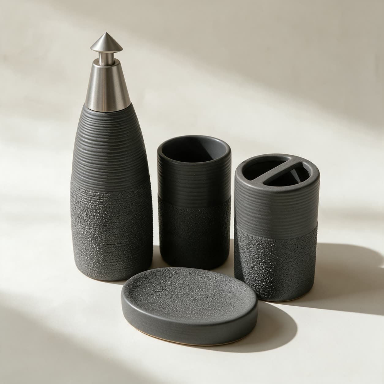 Four-piece charcoal grey bathroom set — a tall ribbed bottle dispenser with a pointed silver pump, a tumbler, a toothbrush holder and a round soap dish — on a warm off-white background