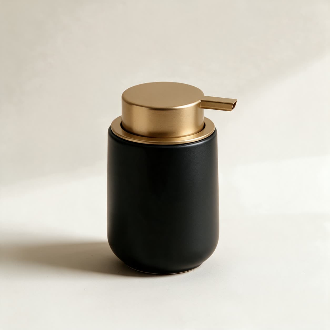 Cylindrical soap dispenser in matte black with a flat brushed gold disc lid and a side press lever, on a warm off-white background