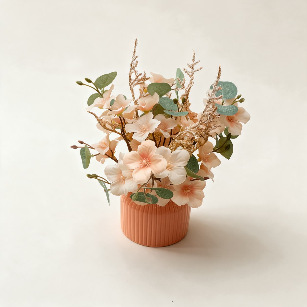 Small artificial arrangement of peach and cream blossoms with eucalyptus leaves and gold sprays in a ribbed terracotta pot, on a warm off-white background