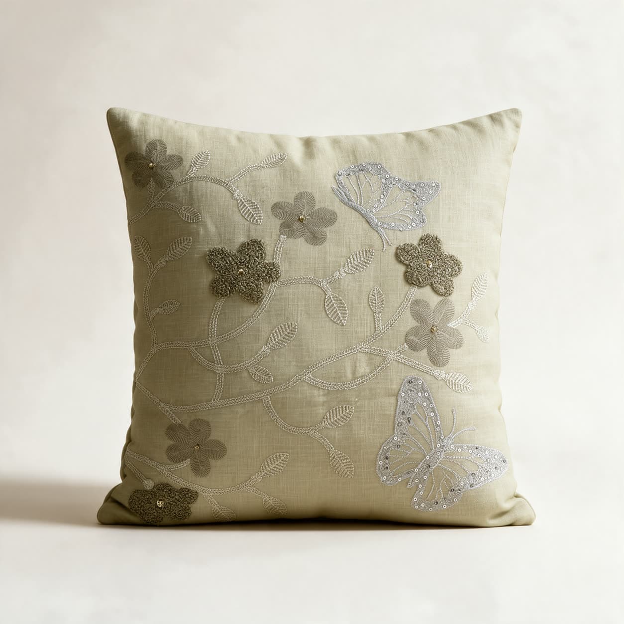 Sage green linen cushion cover hand-embroidered with a silver vine, grey-green flowers with gold bead centres and two silver sequinned butterflies, on a warm off-white background
