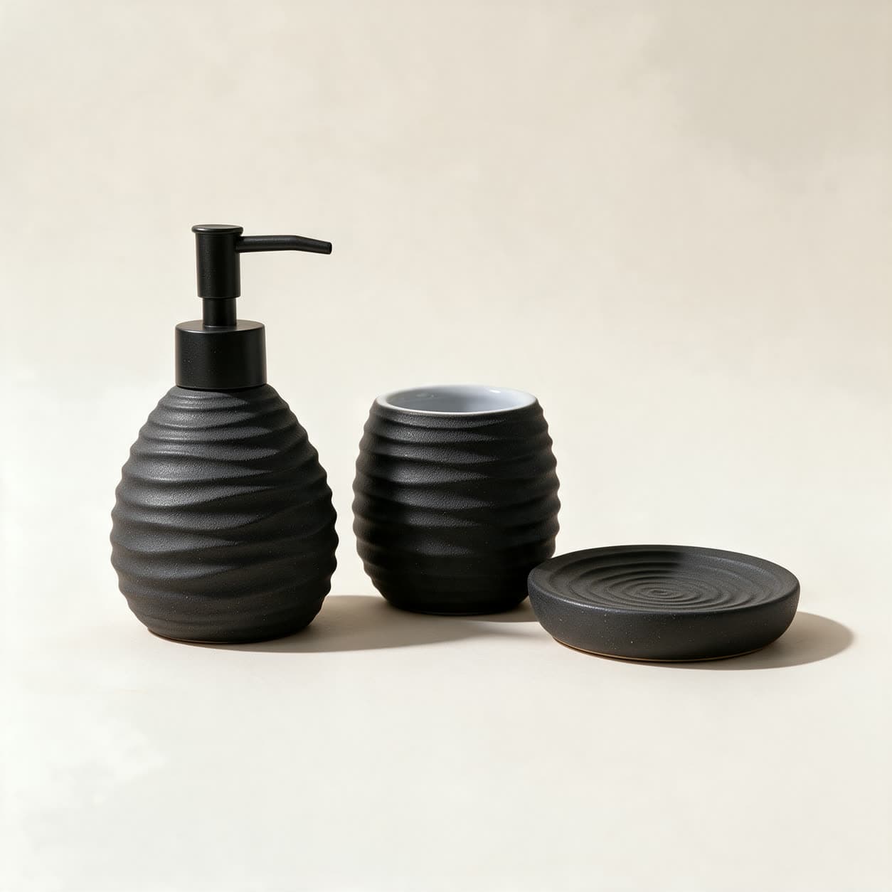 Three-piece matte charcoal ceramic bathroom set — a rippled soap dispenser with a black pump, a matching tumbler and a round soap dish — on a warm off-white background