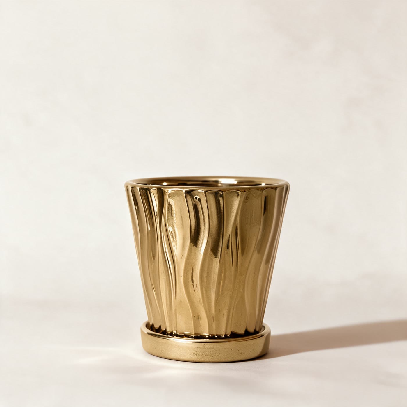 Small mirror-gold ceramic planter moulded in tall wavering flutes, standing alone on its matching gold saucer on a plain warm off-white studio background